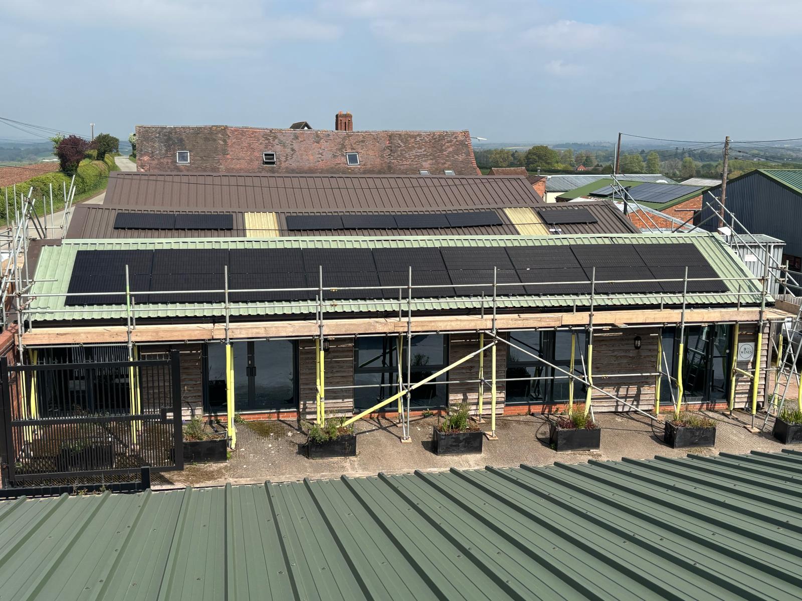 Looking down at the roof of Hard to Find Wines, a newly installed solar array is shown. The panels are flush with the green roof, and the surrounding scaffolding and a bright blue sky suggest a recent, successful installation.