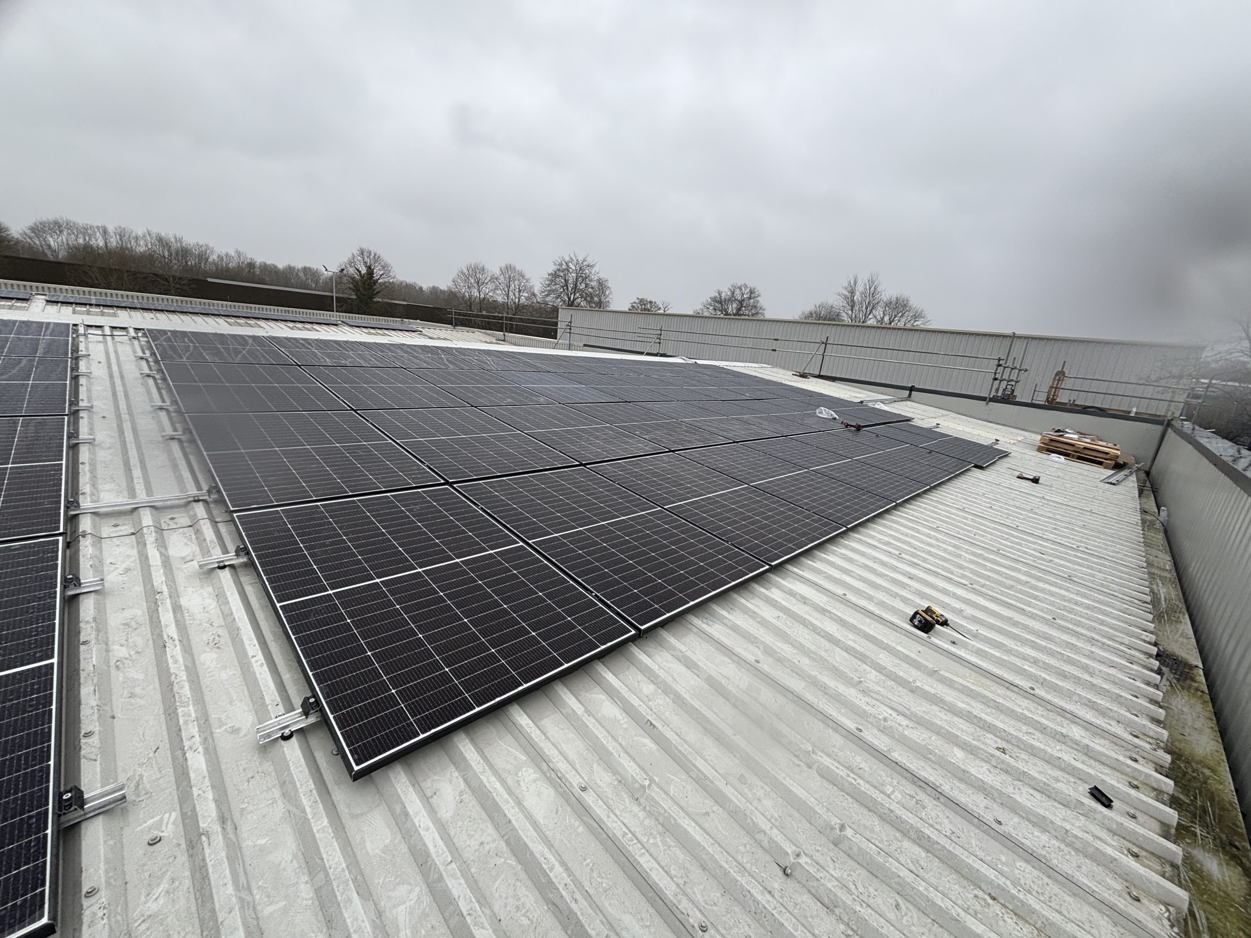 A wide shot shows solar panels mounted on a corrugated metal roof under a gray sky. The panels are neatly arranged in a large rectangle, with trees and a few buildings visible in the background.