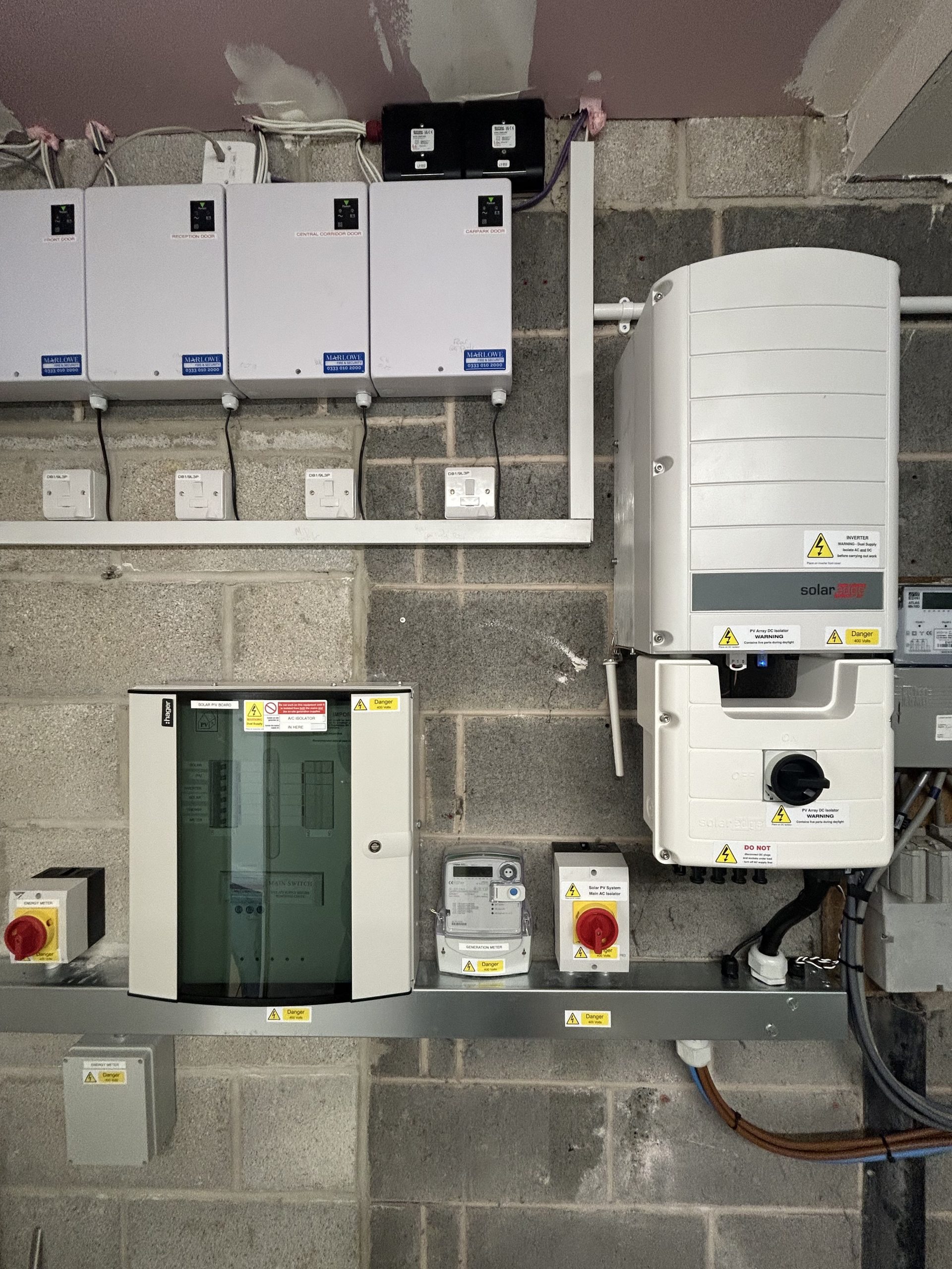 This is an indoor shot of a complete commercial solar panel system. The wall is made of gray cinder blocks and the solar inverter, main fuse box, and other electrical components are mounted on it. The equipment is modern and clean with clear warning labels, and a red power switch is visible to the left and right of the fuse box.