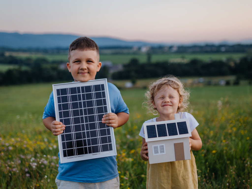 Two children in a field looking happy and smiling holding up solar panel plans for a family home.