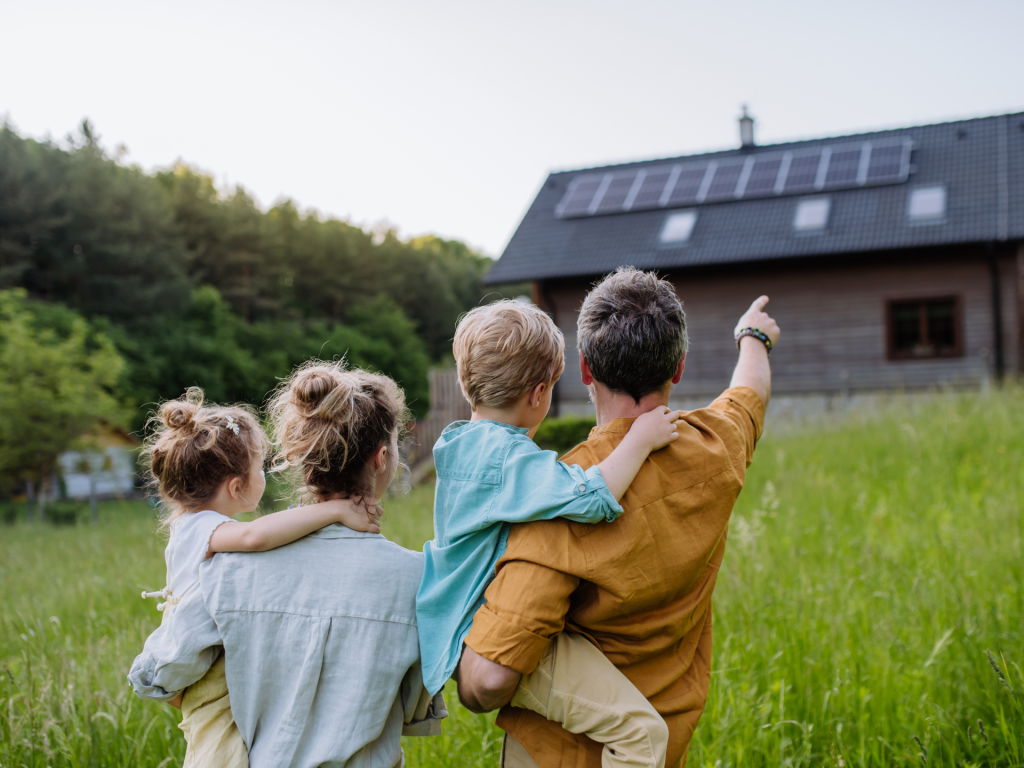 A family of four looking at their house with solar panels installed on the roof. The Dad is pointing at the roof.