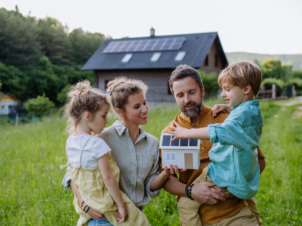A family of four looking at a small mockup house with solar panels on, standing in front of their house with solar panels installed on the roof.